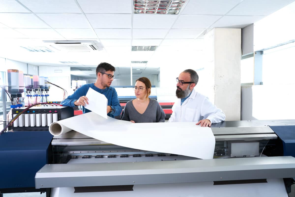 Three people stand in a paper production facility, with inks and other machinery in the background. A man holds a roll of paper while discussing a project with two others.
