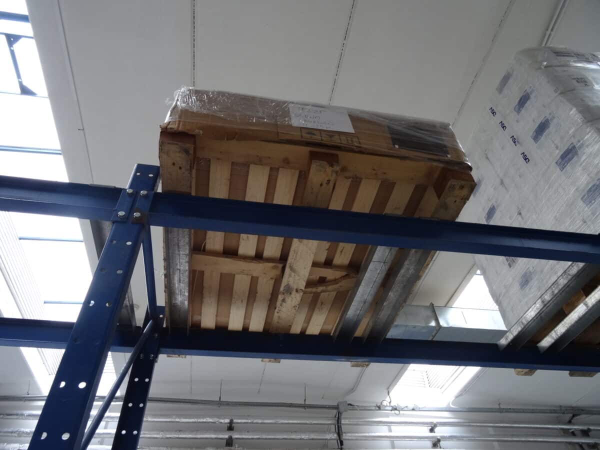 An unstable wooden pallet stacked with corrugated cardboard boxes hangs over the edge of a tall rack in a manufacturing facility, posing safety risks.
