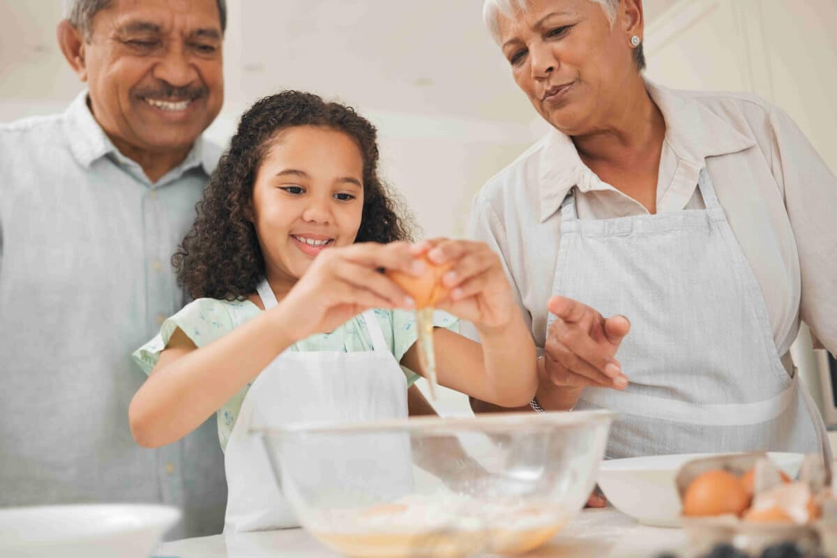 A little girl cracks open a brown egg over a bowl of flour as she prepares a meal with her grandparents in the kitchen.