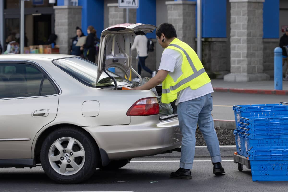 A grocery employee wears a high visibility vest and puts groceries from an online grocery pick up order into a customer's car.