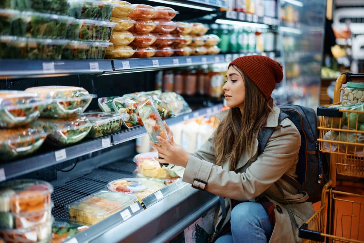 A woman shopping stoops down to observe a container of vegetables in the prepared food aisle of a grocery store.