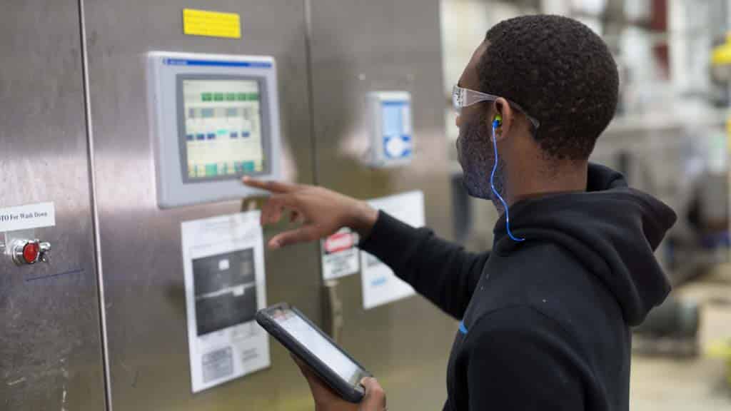 A man wearing ear plugs and safety eyeglasses touches a monitor displayed on a stainless-steel door, using technology to track the functionality of machinery in an industrial facility.
