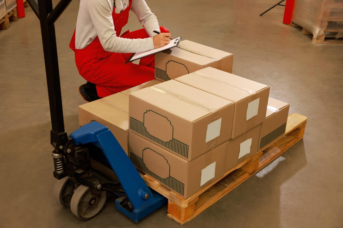 A retail worker holding a clipboard of documents and a pen crouches down in a store backroom to manually check the inventory of cardboard boxes stacked on a dolly pallet. This manual data collation process is time-consuming and has the potential for human error.